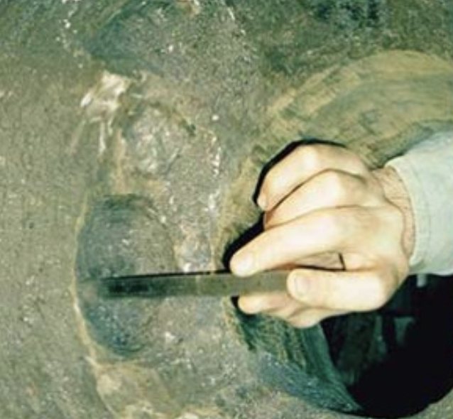 A gloved hand probing a corrosion cavity in a digester wall with a flat scraper, showing how far the void undercuts the surrounding surface.